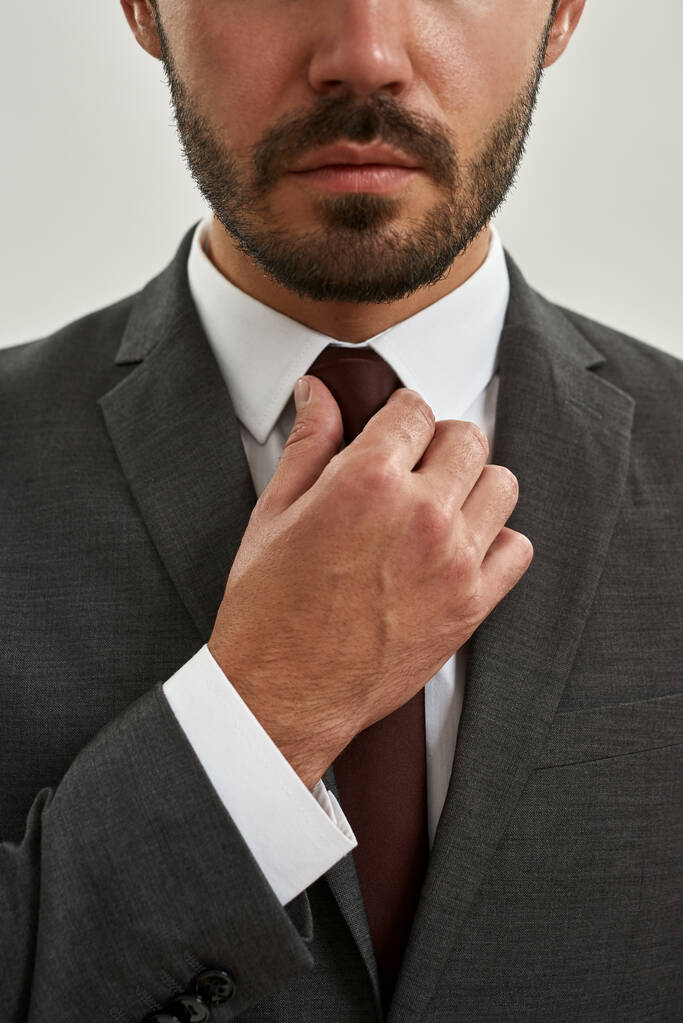 Częściowo poważny brodaty przedsiębiorca lub szef firmy wyprostować krawat garnituru. Pojęcie nowoczesnego, odnoszącego sukcesy męskiego stylu życia. Odizolowany na białym tle. Zdjęcia studyjne - Zdjęcie, obraz