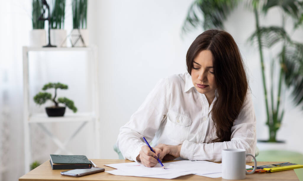 Een donkerharig creatief enthousiast meisje in een keurig wit stijlvol shirt zit aan een bureau en tekent haar notitieboekje of maakt notities in haar dagboek, in haar gezellige lichte kamer - Foto, afbeelding