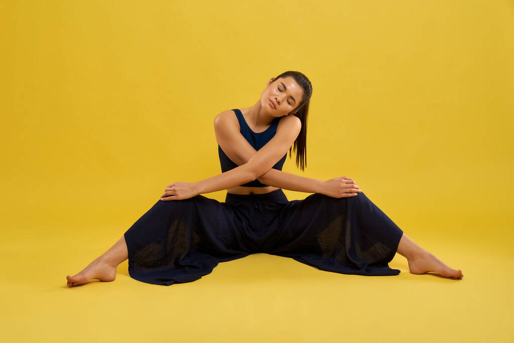 Молодая женщина делает упражнения на растяжку, сидит с широко расставленными ногами в помещении. Вид спереди расслабленной девушки в черном, со скрещенными руками на коленях, изолированной на оранжевом фоне. Концепция медитации. - Фото, изображение