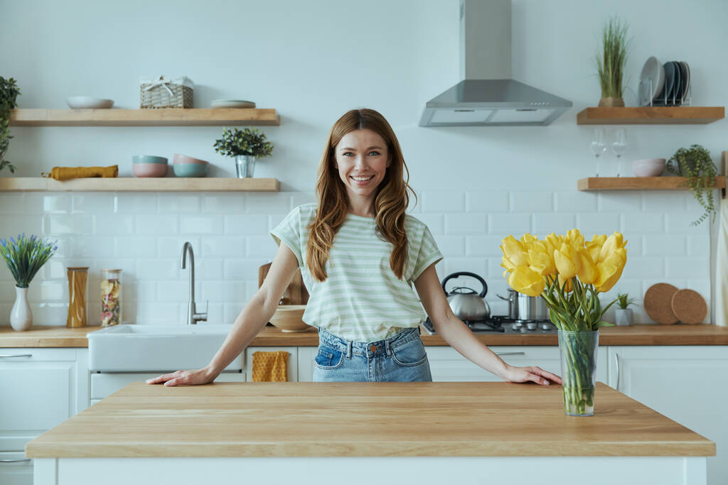 Beautiful young woman leaning at the kitchen desk and smiling - Photo, Image