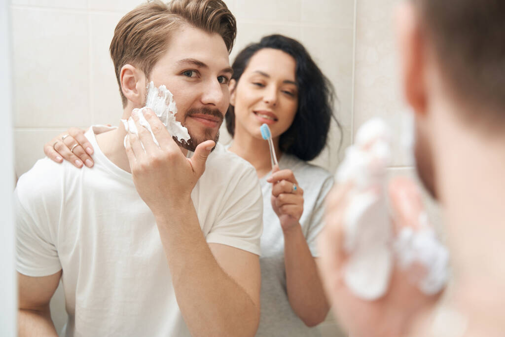 Γυναίκα με οδοντόβουρτσα στο χέρι παρατηρώντας αρσενικό σύντροφό της εφαρμογή αφρό ξυρίσματος στο πρόσωπο - Φωτογραφία, εικόνα