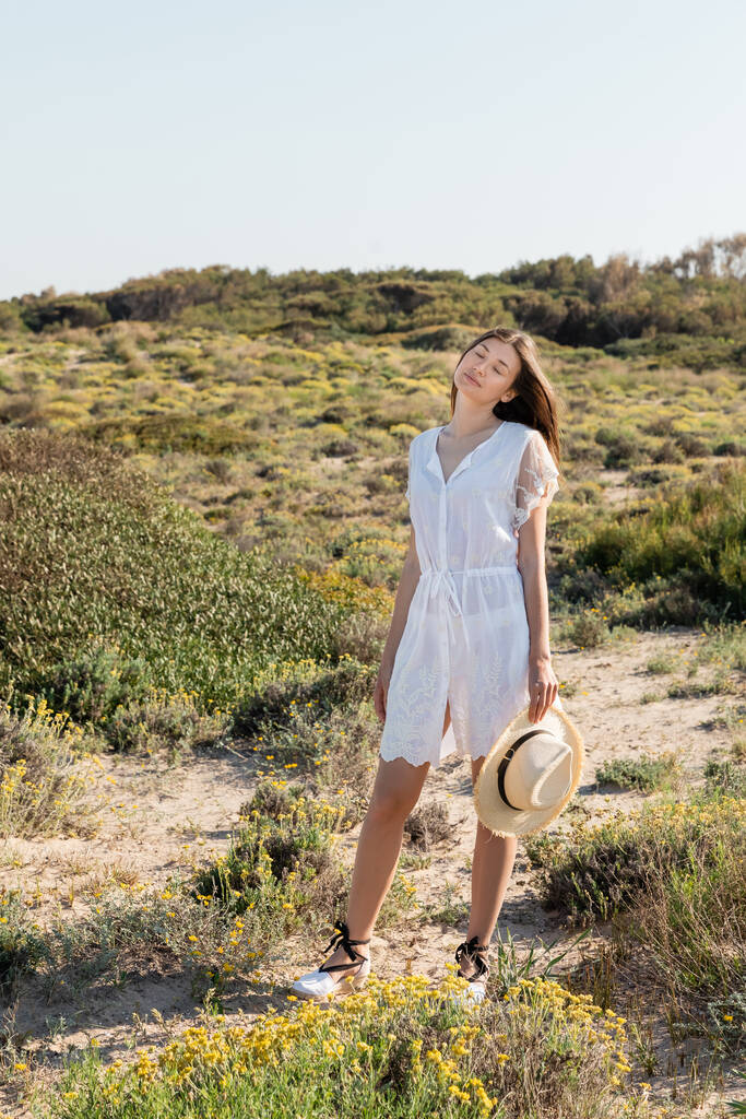 Jovem mulher de vestido fechando os olhos e segurando chapéu de palha enquanto estava na praia  - Foto, Imagem