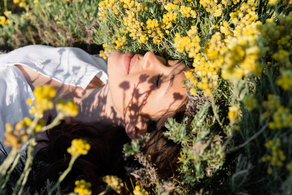 Młoda kobieta zamyka oczy leżąc na łące z kwiatami  - Zdjęcie, obraz