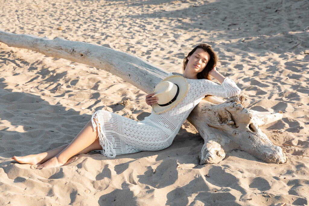 Молодая женщина в платье, держащая шляпу от солнца, отдыхая возле деревянного бревна на песчаном пляже  - Фото, изображение