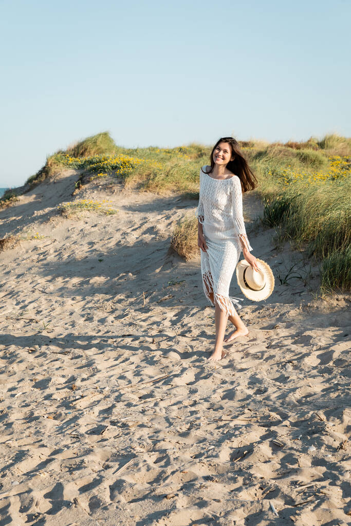 Mulher morena positiva no vestido segurando chapéu de palha enquanto caminhava na praia de areia  - Foto, Imagem