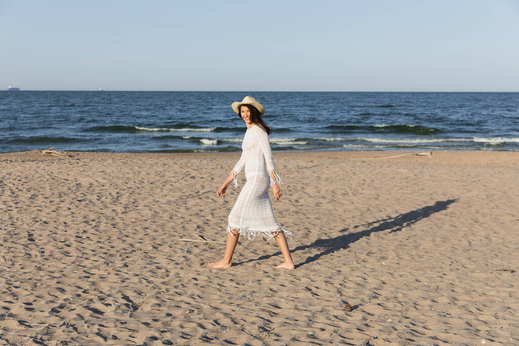 Femme heureuse en robe et chapeau de soleil regardant la caméra tout en marchant sur la plage près de la mer  - Photo, image