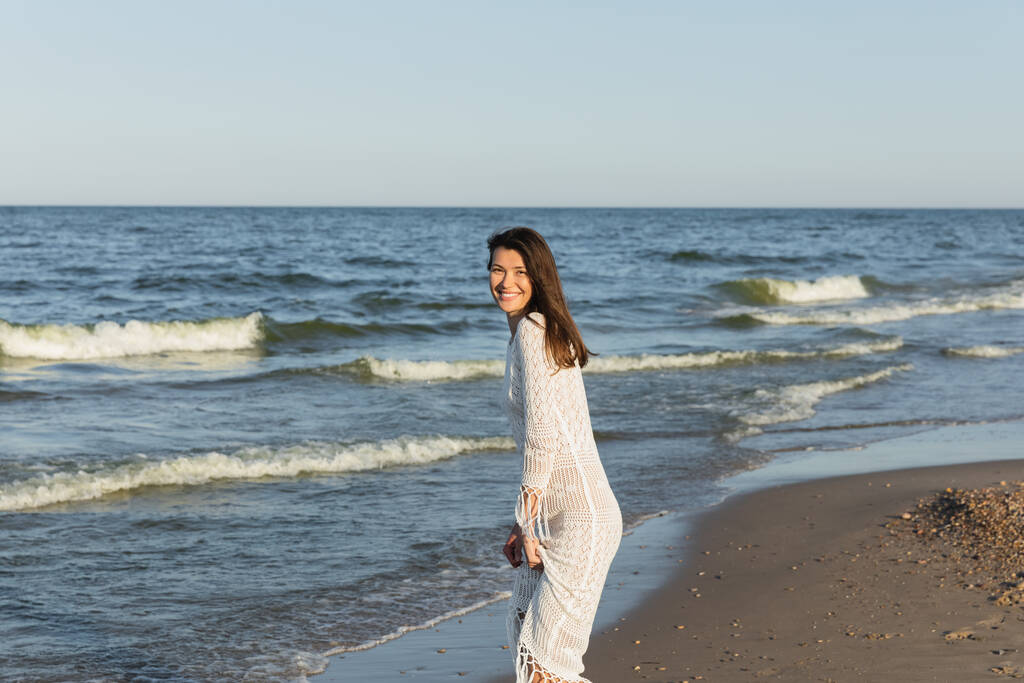 Όμορφη γυναίκα με φόρεμα χαμογελά στην κάμερα κοντά στη θάλασσα στην παραλία  - Φωτογραφία, εικόνα