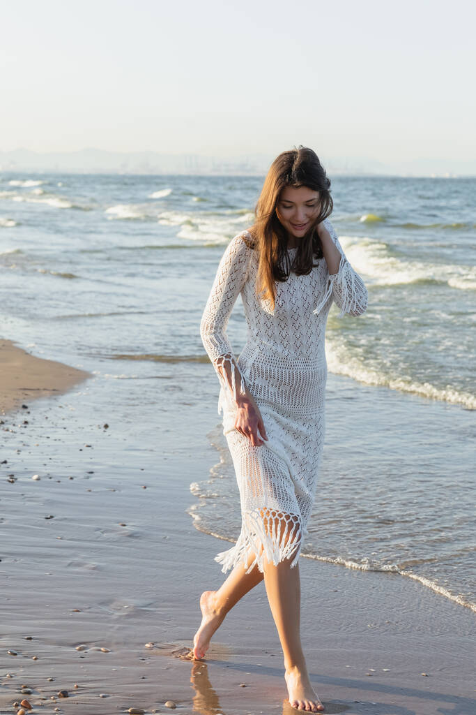 Позитивна молода жінка в літній сукні, що йде на мокрий пісок на пляжі
  - Фото, зображення