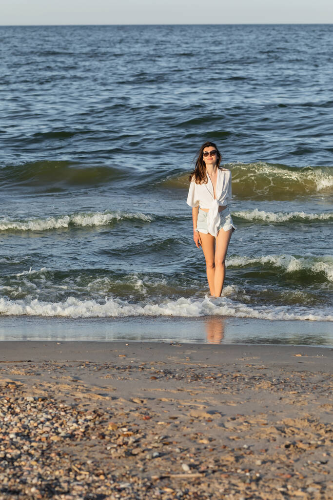 Mujer joven en gafas de sol de pie en el agua de mar cerca de la playa  - Foto, imagen