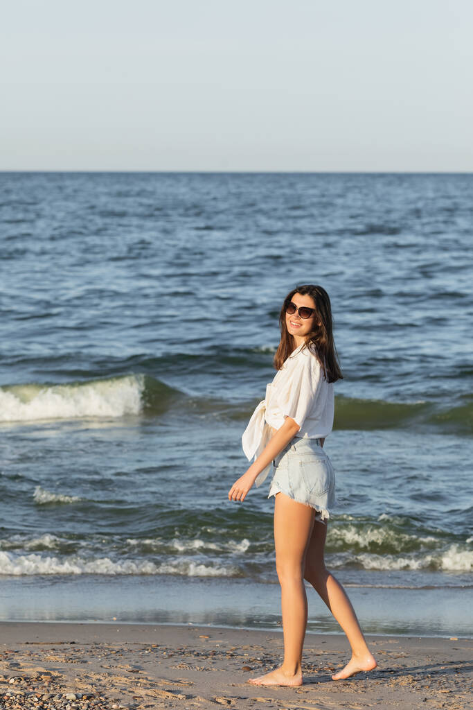 Позитивная молодая женщина в солнечных очках ходит по мокрому песку на пляже  - Фото, изображение