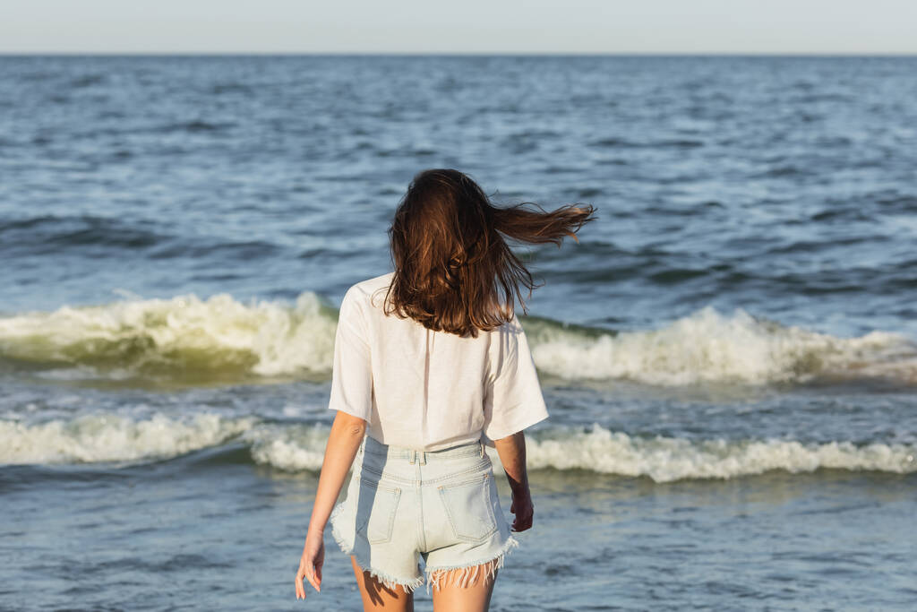 Vista trasera de mujer morena en pantalones cortos de mezclilla de pie cerca del mar borroso  - Foto, Imagen