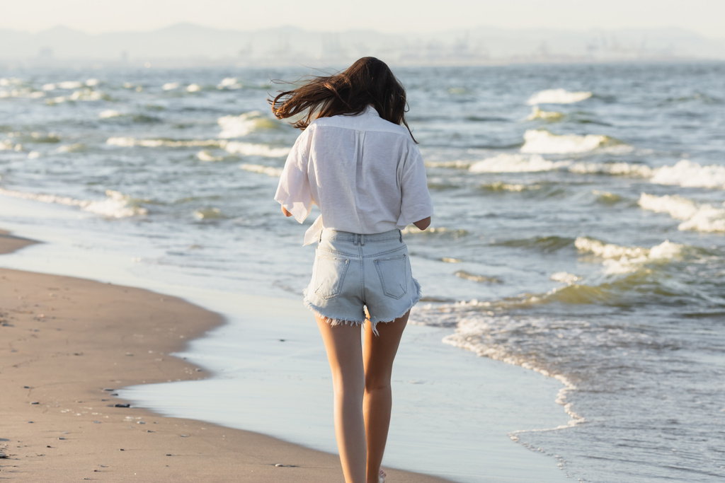 Вид сзади женщины в белой рубашке и джинсовых шортах, прогуливающейся у моря на пляже  - Фото, изображение