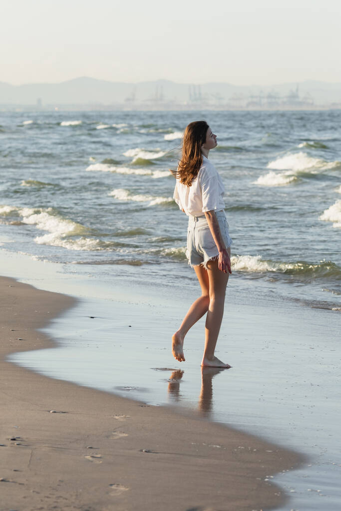 Πλευρική άποψη της νεαρής γυναίκας με σορτς στέκεται σε υγρή άμμο κοντά στη θάλασσα  - Φωτογραφία, εικόνα