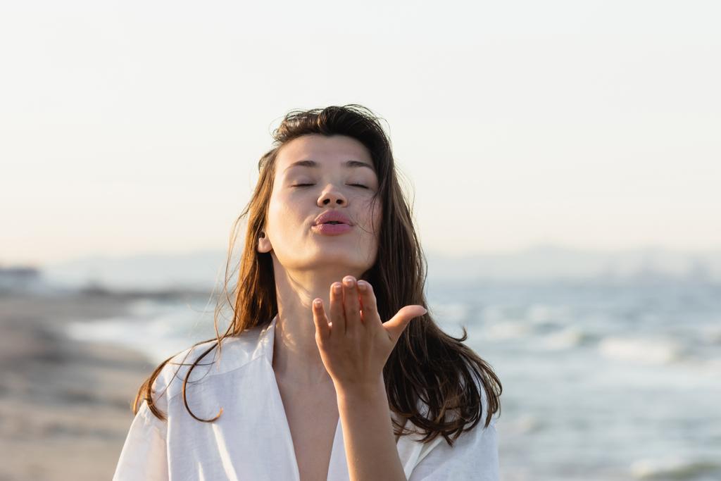 Brunette vrouw in wit shirt blazen lucht kus op wazig strand  - Foto, afbeelding