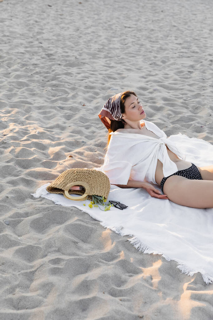 Γυναίκα με μαγιό και πουκάμισο αναπαύεται κοντά στην τσάντα και γυαλιά ηλίου στην παραλία  - Φωτογραφία, εικόνα