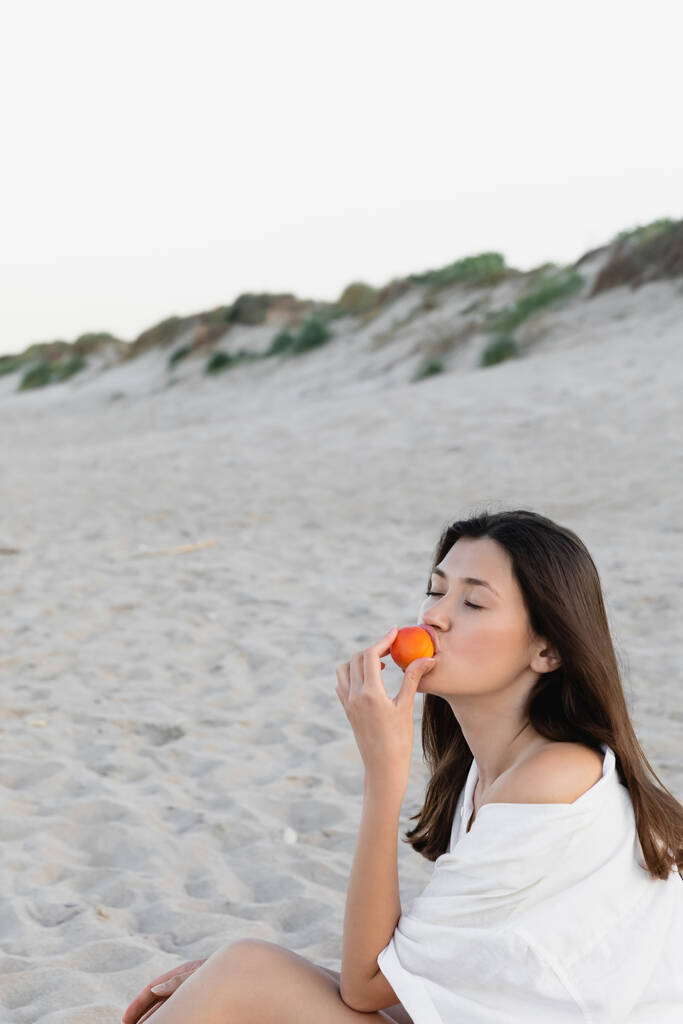 Mulher morena na camisa comendo damasco na praia  - Foto, Imagem