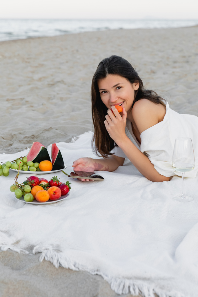 Mujer sonriente sosteniendo albaricoque y teléfono inteligente cerca de vino y frutas en la playa  - Foto, Imagen