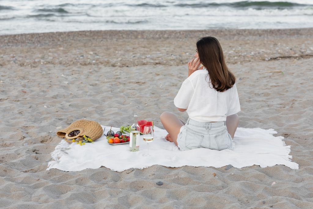Woman talking on smartphone near wine and fruits on beach  - Photo, Image