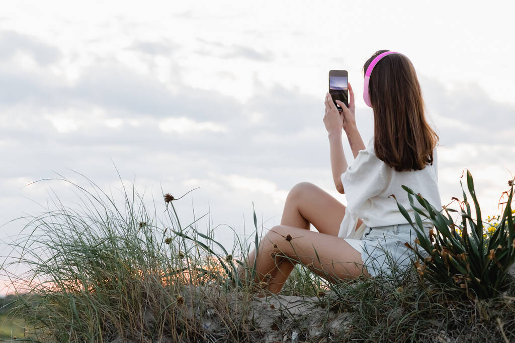 Takaisin näkymä nainen kuulokkeet ottaa valokuvan älypuhelin rannalla illalla  - Valokuva, kuva
