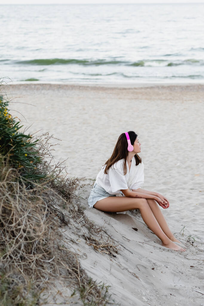 Πλευρική άποψη της νεαρής γυναίκας με πουκάμισο ακούγοντας μουσική σε ακουστικά στην παραλία κοντά στη θάλασσα  - Φωτογραφία, εικόνα