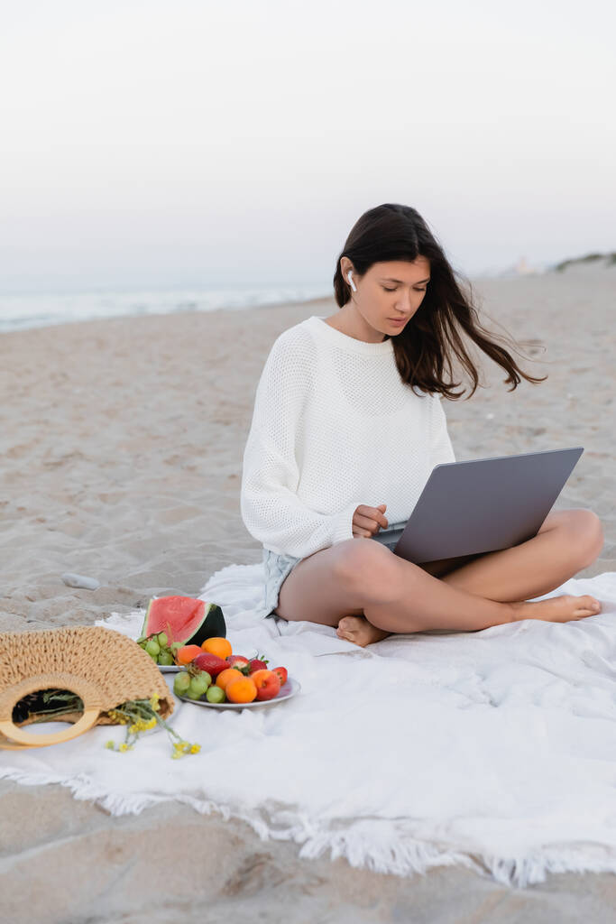 Жінка в навушниках використовує ноутбук біля фруктів на ковдрі.  - Фото, зображення
