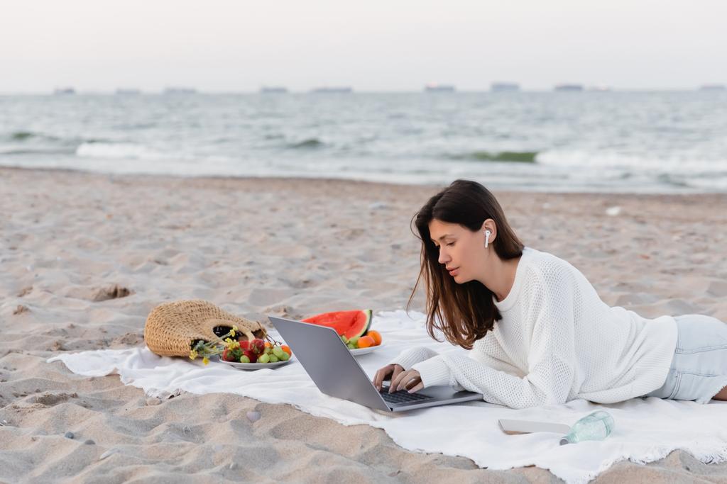 Frau mit Kopfhörer benutzt Laptop in der Nähe von Smartphone und Obst auf Decke am Strand  - Foto, Bild