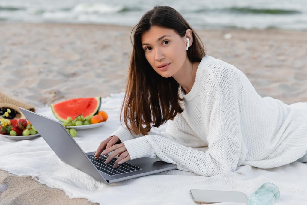 Young freelancer in earphone using laptop near blurred fruits and smartphone on beach  - Photo, Image