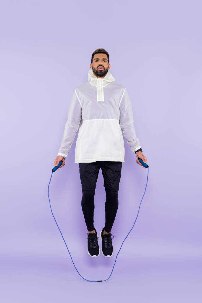longueur totale de sportif afro-américain en sportswear sautant avec corde à sauter sur violet - Photo, image