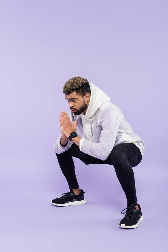 longitud completa de barbudo afroamericano deportista en ropa deportiva que trabaja con las manos de oración en púrpura  - Foto, Imagen