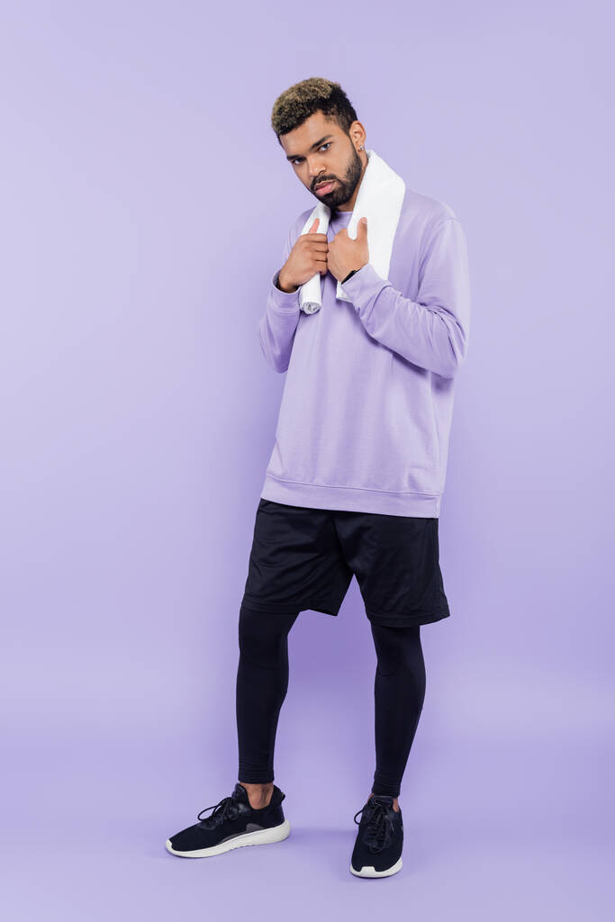 full length of bearded african american man in sweater holding white towel and looking at camera on purple  - Photo, Image