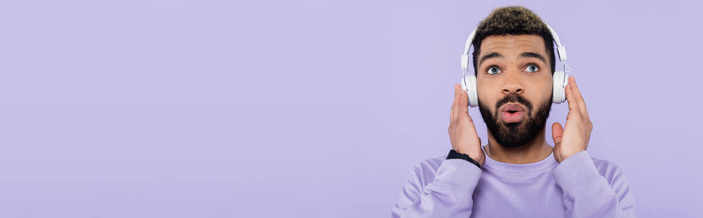 homme afro-américain surpris et barbu dans des écouteurs sans fil isolés sur violet, bannière - Photo, image