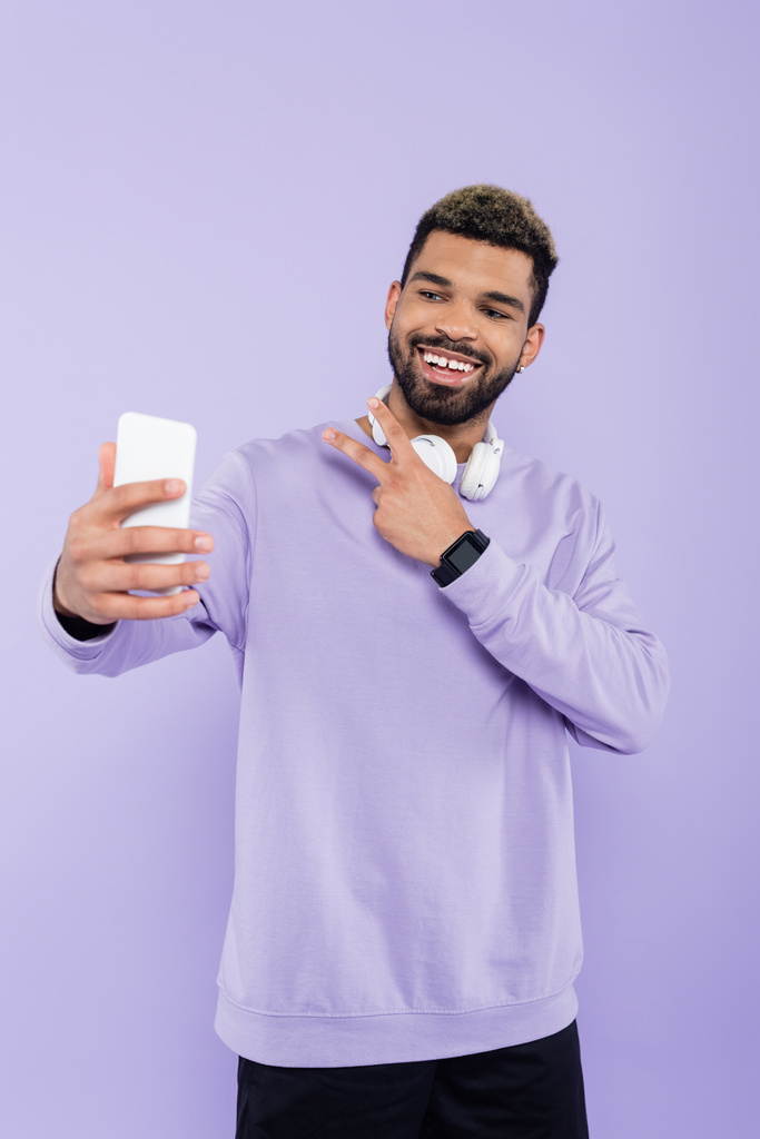 šťastný africký Američan s bezdrátovými sluchátky, přičemž selfie při zobrazení mírové znamení izolované na fialové - Fotografie, Obrázek