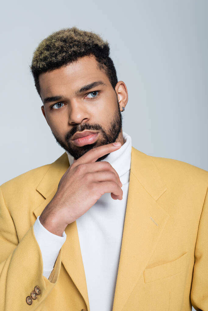 hombre afroamericano pensativo en elegante chaqueta amarilla posando aislado en gris - Foto, imagen