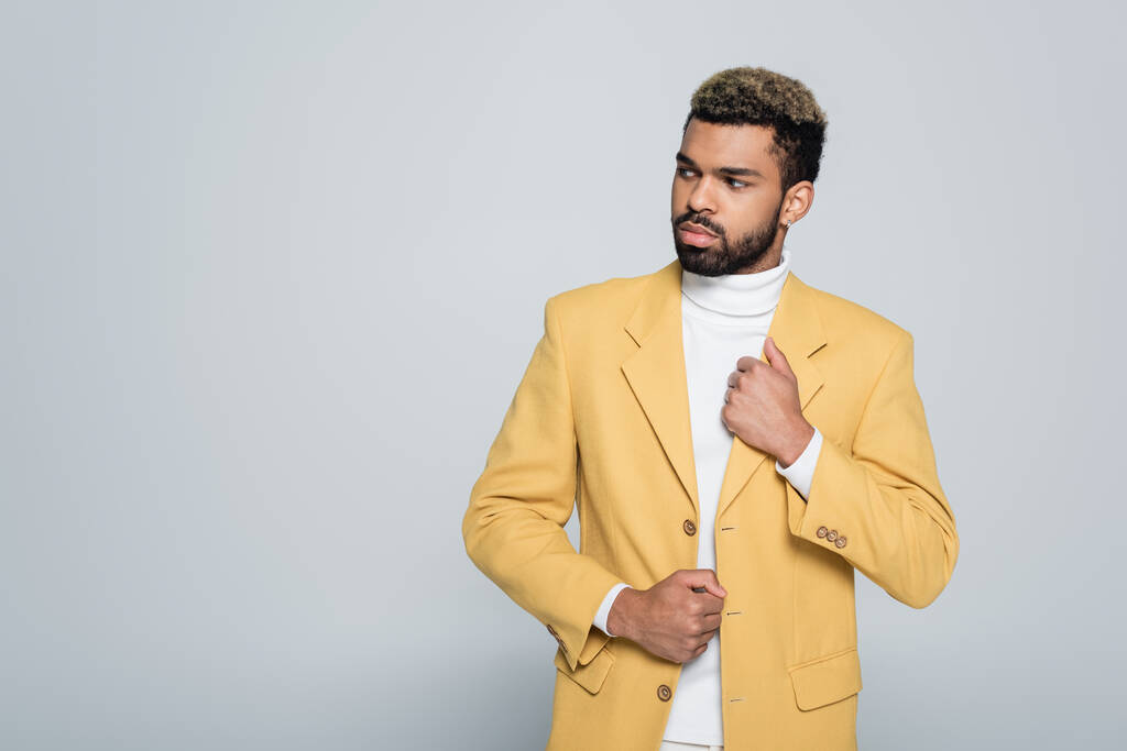 hombre afroamericano con estilo en chaqueta amarilla mirando hacia otro lado aislado en gris - Foto, Imagen