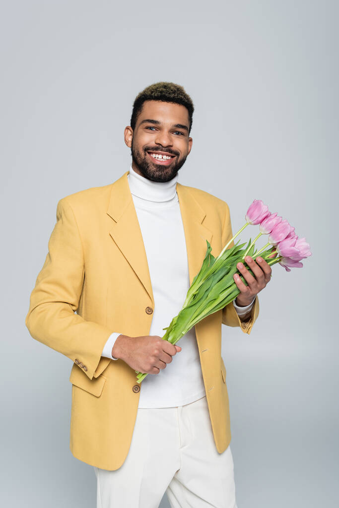 happy african american man in stylish outfit holding bouquet of pink tulips isolated on grey  - Photo, Image