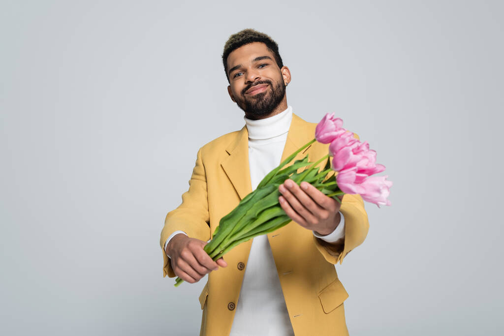 potěšen africký Američan ve stylovém oblečení drží kytice růžových tulipánů izolovaných na šedé  - Fotografie, Obrázek