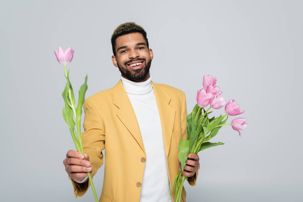 joyful african american man in stylish outfit holding bouquet of pink tulips isolated on grey  - Photo, Image