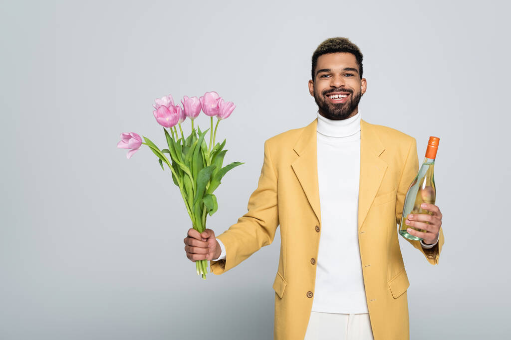 счастливый африканский американец в стильном наряде с букетом розовых тюльпанов, изолированных на сером  - Фото, изображение