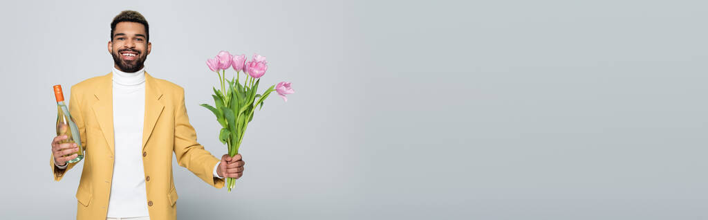 pozitivní africký Američan ve stylovém oblečení držící kytici růžových tulipánů izolovaných na šedé, banner - Fotografie, Obrázek