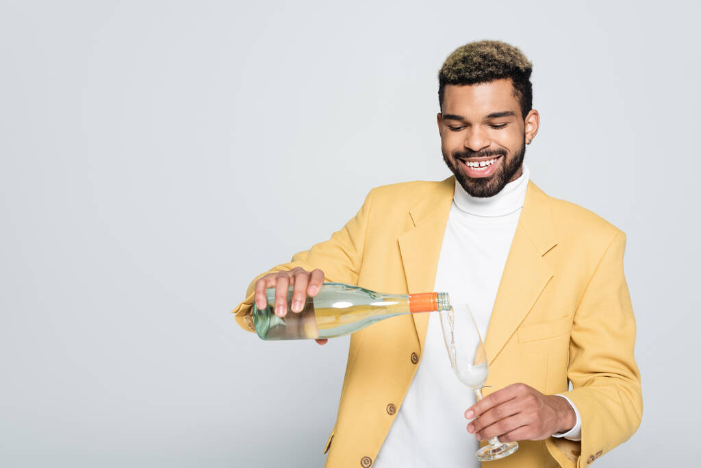 glücklicher afrikanisch-amerikanischer Mann in stylischem Outfit mit Flasche und Champagner im Glas isoliert auf grau  - Foto, Bild