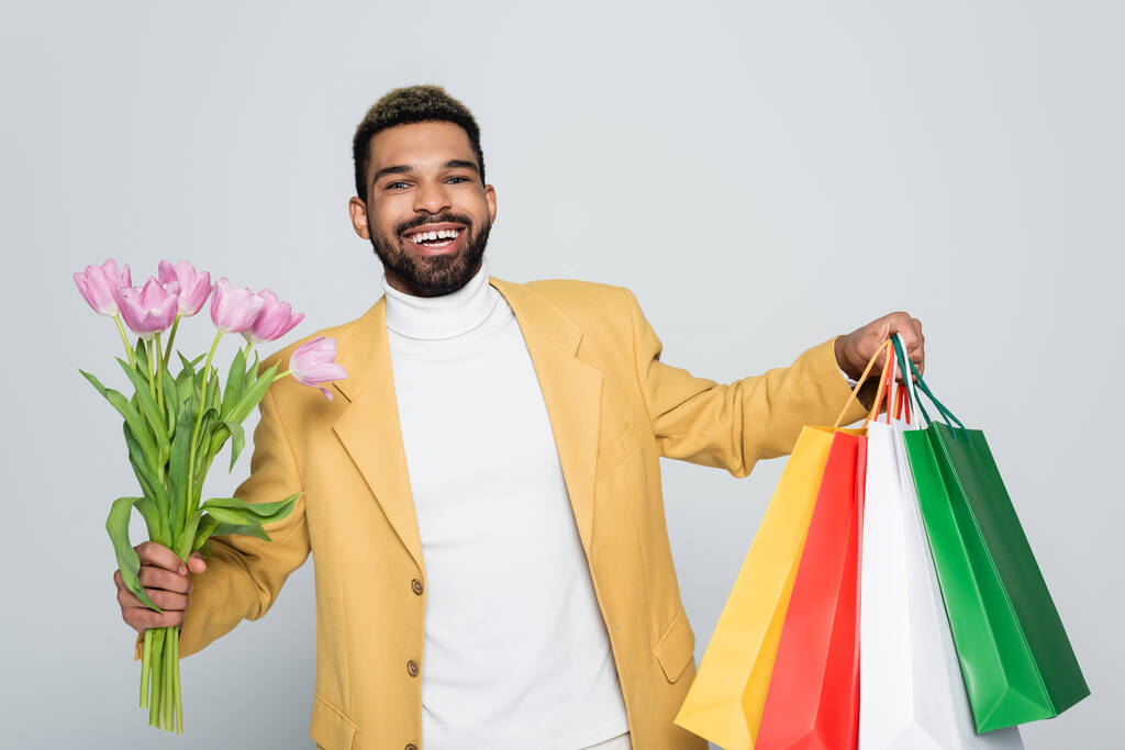allegro uomo afroamericano in giacca gialla e collo polo con tulipani rosa e shopping bag isolati su grigio  - Foto, immagini