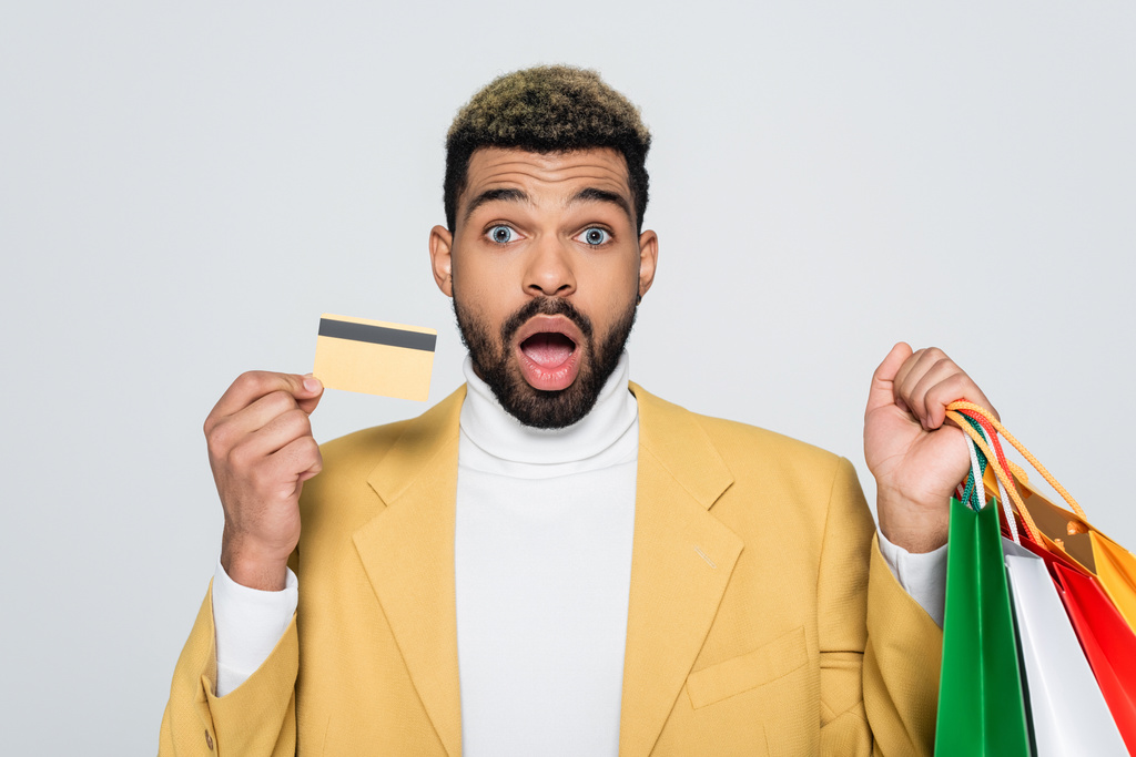 meglepett afro-amerikai férfi sárga blézerben kezében bevásárló táskák és hitelkártya elszigetelt szürke  - Fotó, kép