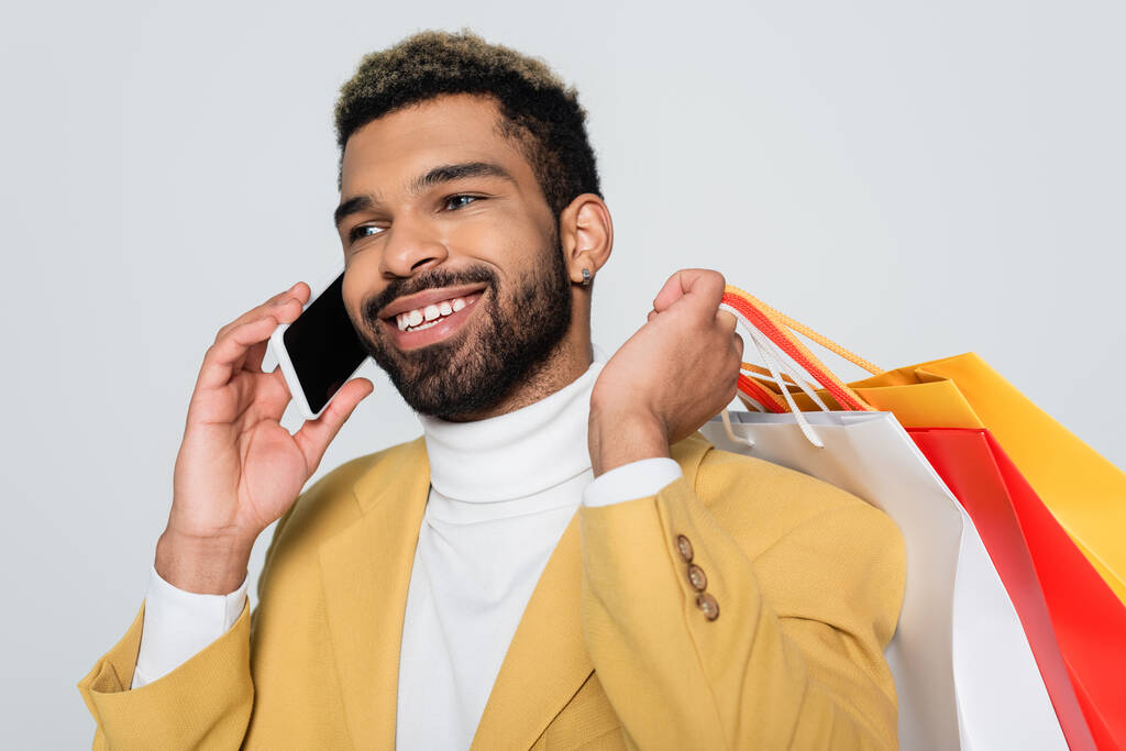 alegre hombre afroamericano en blazer amarillo y cuello polo sosteniendo bolsas de compras y hablando por teléfono celular aislado en gris  - Foto, Imagen