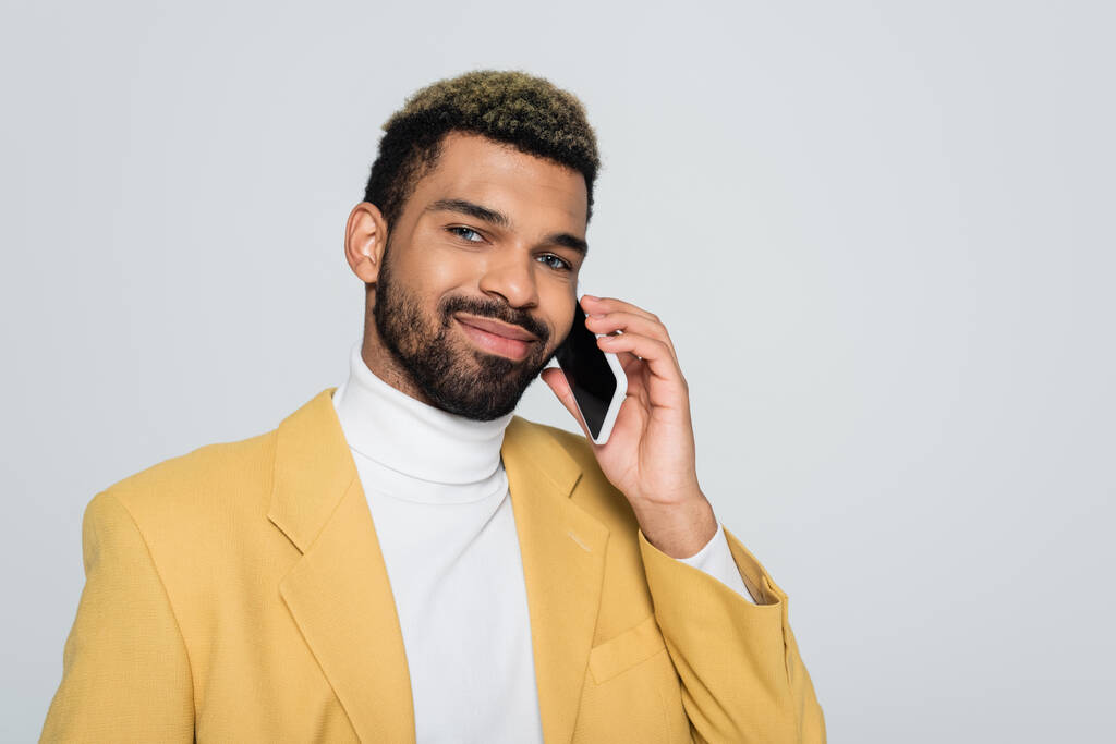 hombre americano africano alegre en chaqueta amarilla hablando en teléfono inteligente aislado en gris  - Foto, imagen
