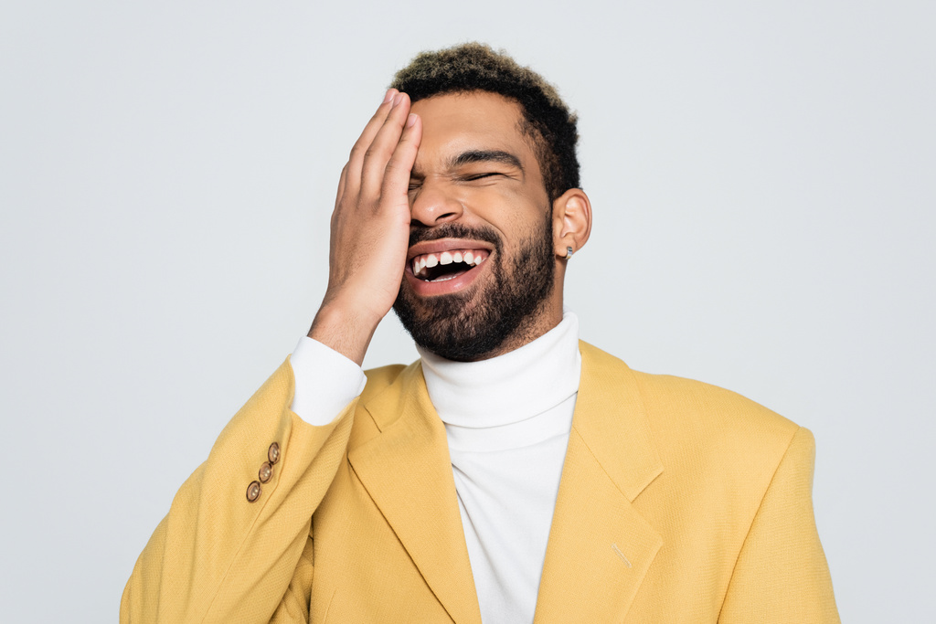 幸せなアフリカ系アメリカ人男性でパステルイエローのブレザーで目を閉じて笑ってグレーで隔離された  - 写真・画像