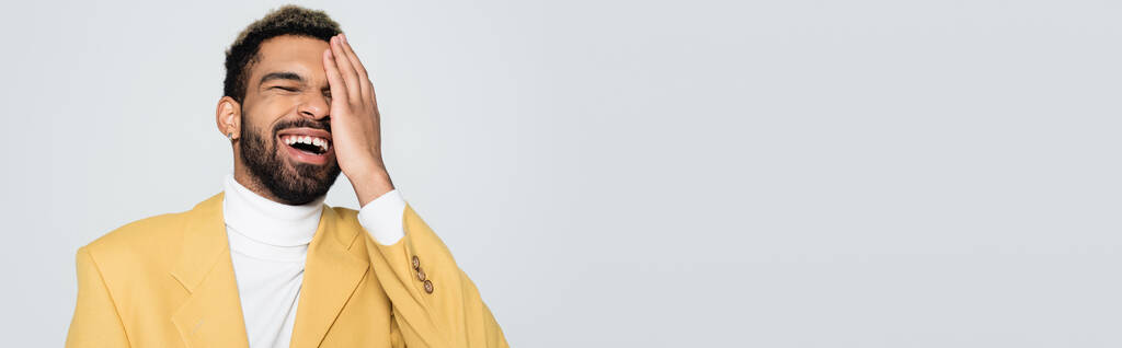 Pastel sarısı ceketli mutlu Afro-Amerikan adam gözlerini kapatıyor ve gri pankartta tek başına gülüyor. - Fotoğraf, Görsel