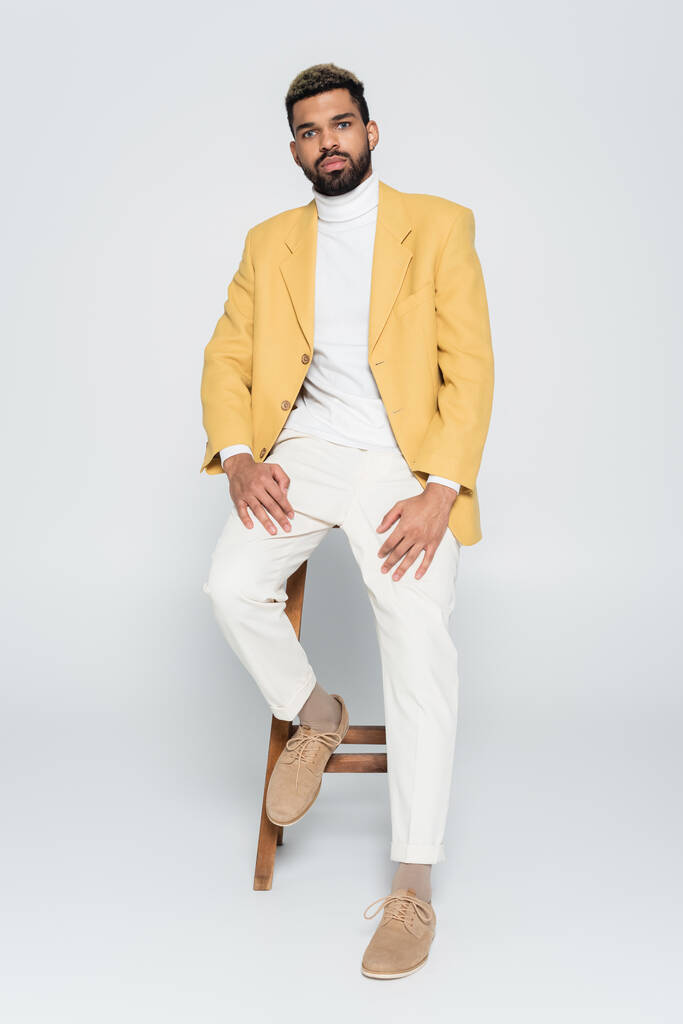 volle Länge des bärtigen afrikanisch-amerikanischen Mann in stilvollem Outfit sitzt auf einem hölzernen Hochstuhl auf grau  - Foto, Bild