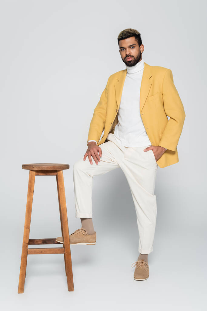 toute la longueur de barbu homme afro-américain en tenue élégante debout avec la main dans la poche près de chaise haute en bois sur gris  - Photo, image