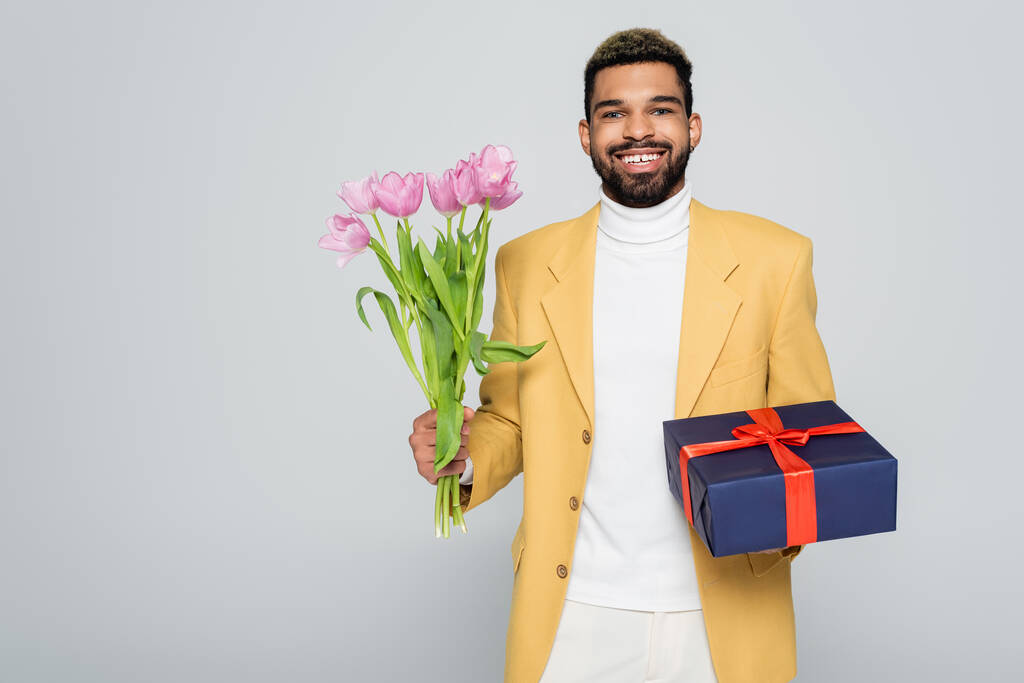 happy african american man in yellow stylish blazer holding wrapped present and tulips isolated on grey  - Photo, Image