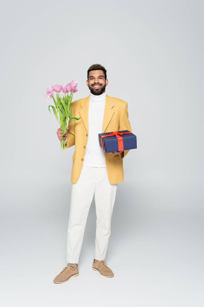 longitud completa de hombre afroamericano feliz en chaqueta amarilla sosteniendo regalo envuelto y tulipanes en gris  - Foto, imagen